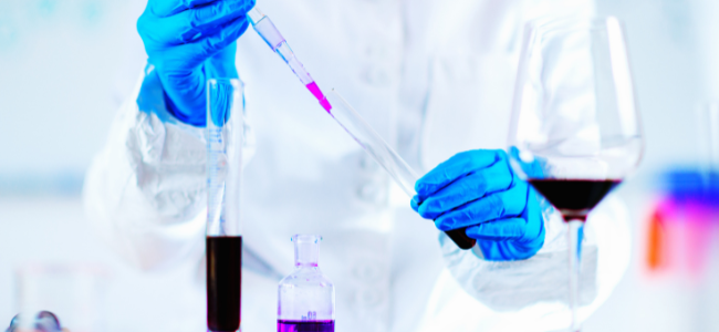 Científico en laboratorio realizando una valoración de sulfuroso, utilizando una pipeta y probetas, con una copa de vino al lado